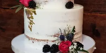 A two-tier semi-naked wedding cake with white frosting, gold leaf accents, and fresh floral and berry decorations on a green cake stand.