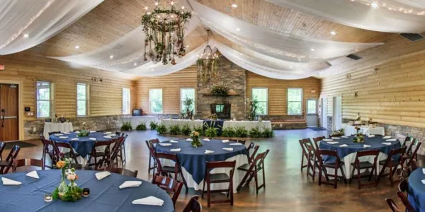 Interior view of a wedding reception hall featuring wood-paneled walls, a stone fireplace, chandeliers, and round tables set with blue tablecloths.