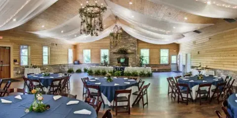 Interior view of a wedding reception hall featuring wood-paneled walls, a stone fireplace, chandeliers, and round tables set with blue tablecloths.