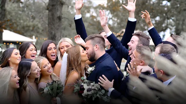 Bride and groom surrounded by a cheering wedding party with raised hands at an outdoor wedding.