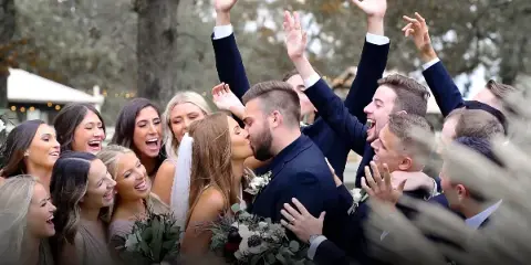 Bride and groom surrounded by a cheering wedding party with raised hands at an outdoor wedding.