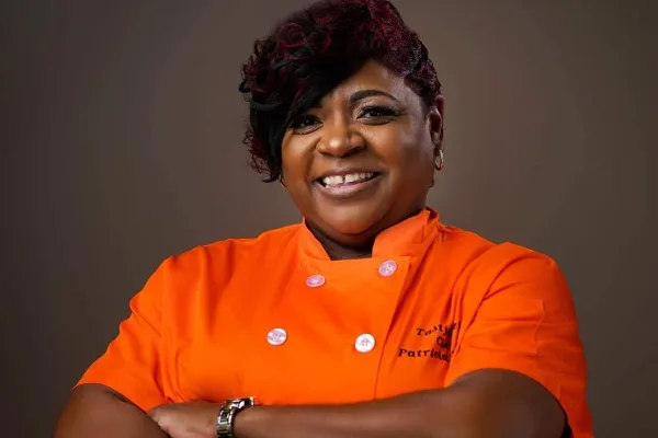 Portrait of a smiling woman in an orange chef's coat with 'Tasty Tee's' embroidered on the chest.