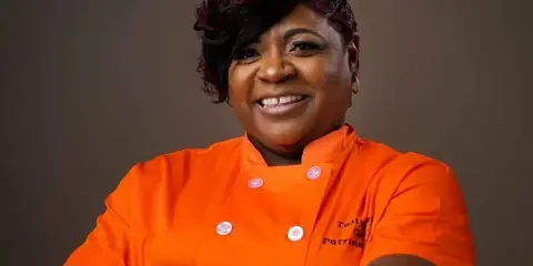 Portrait of a smiling woman in an orange chef's coat with 'Tasty Tee's' embroidered on the chest.