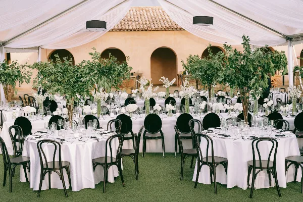 A wedding reception setup under a white tent with round tables, white tablecloths, black chairs, and floral arrangements.