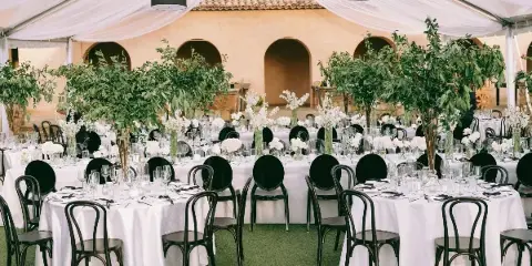 A wedding reception setup under a white tent with round tables, white tablecloths, black chairs, and floral arrangements.