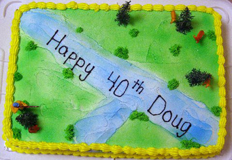 A rectangular sheet cake decorated with a green landscape, a blue river, and the text 'Happy 40th Doug'.