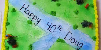 A rectangular sheet cake decorated with a green landscape, a blue river, and the text 'Happy 40th Doug'.
