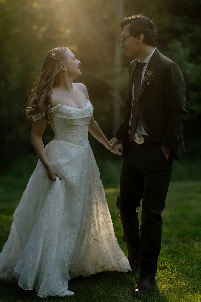 A bride and groom holding hands and smiling at each other in a sunlit outdoor setting.