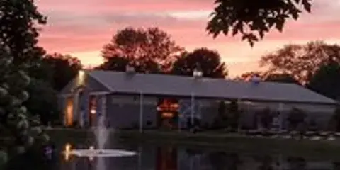 A large barn-style wedding venue building reflected in a pond at sunset.