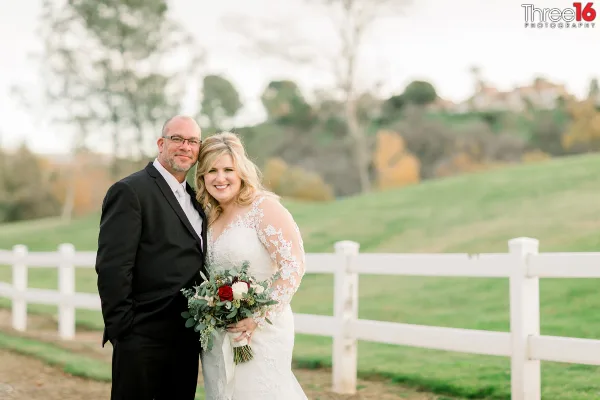 A professional wedding photograph of a bride and groom standing together outdoors near a white fence.