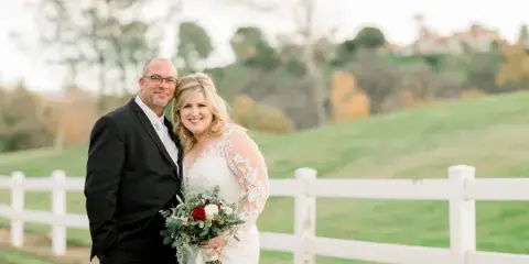 A professional wedding photograph of a bride and groom standing together outdoors near a white fence.