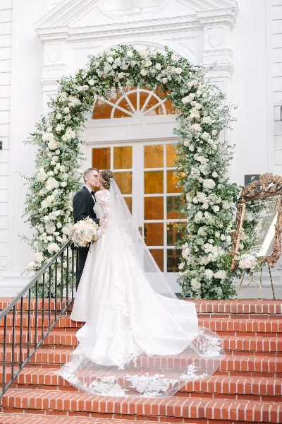A bride and groom kissing on brick steps in front of a large, white floral archway at a wedding venue.