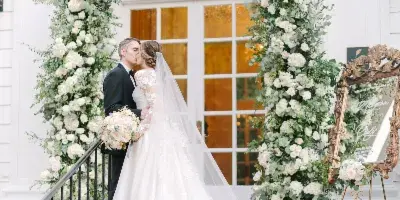 A bride and groom kissing on brick steps in front of a large, white floral archway at a wedding venue.