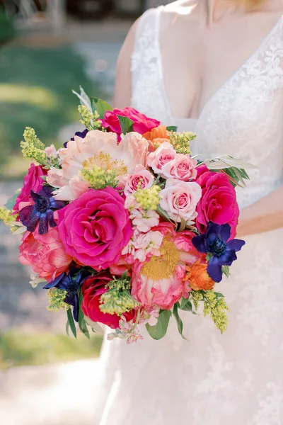 A close-up photograph of a bride in a white lace dress holding a colorful bouquet of pink, orange, and purple flowers.