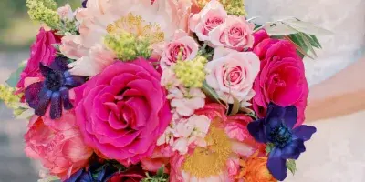 A close-up photograph of a bride in a white lace dress holding a colorful bouquet of pink, orange, and purple flowers.