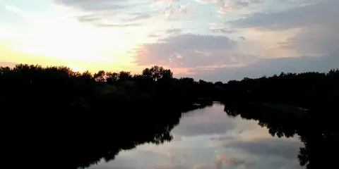A calm river reflecting a sunset sky, bordered by dark silhouettes of trees and vegetation.