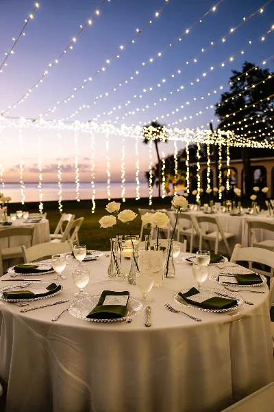 A wedding reception table set with white linens, green napkins, and white roses under a canopy of string lights at sunset.