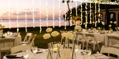 A wedding reception table set with white linens, green napkins, and white roses under a canopy of string lights at sunset.