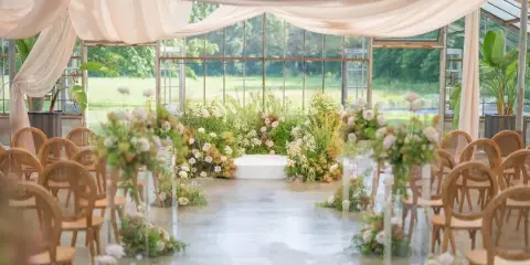 A wedding ceremony setup in a glass-walled venue featuring a floral altar, white drapery, and rows of wooden chairs.