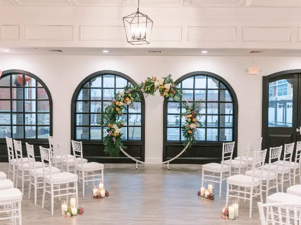 Interior view of a wedding venue with white chairs, a floral arch, and large arched windows.