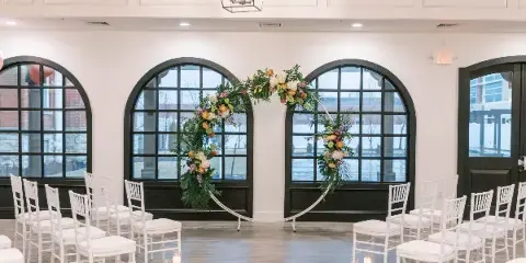 Interior view of a wedding venue with white chairs, a floral arch, and large arched windows.