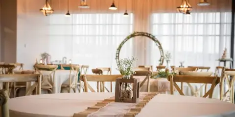 A wedding reception hall with round tables, wooden chairs, and a floral arch in the background.