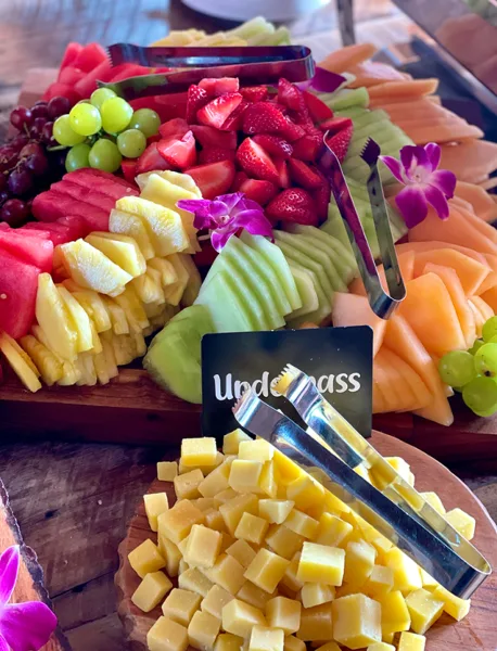 A catering spread featuring a wooden board with sliced melons, strawberries, grapes, and pineapple, alongside a bowl of cubed cheese and serving tongs.