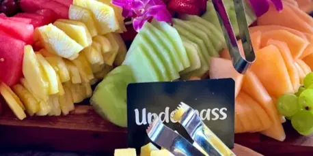 A catering spread featuring a wooden board with sliced melons, strawberries, grapes, and pineapple, alongside a bowl of cubed cheese and serving tongs.
