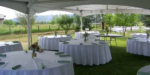 An outdoor event setup featuring a large white tent and several round tables with white tablecloths and green napkins.
