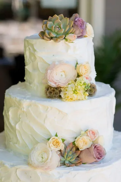 A two-tier white wedding cake decorated with pastel roses, ranunculus, and succulents.