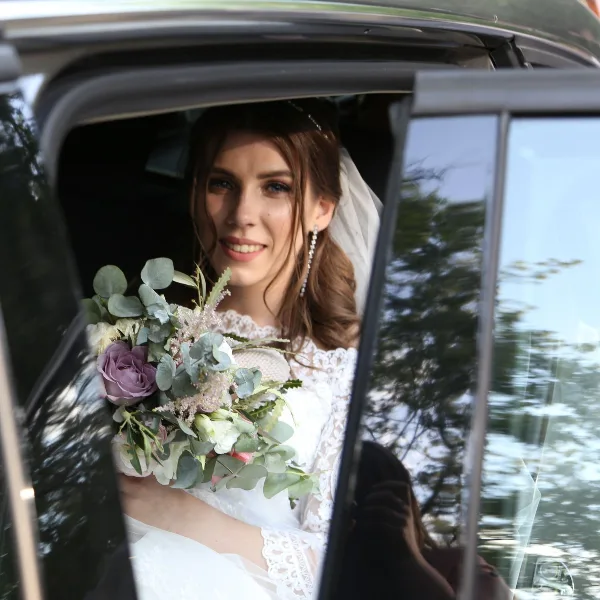A bride in a white lace wedding dress and veil holding a floral bouquet while seated in a car.