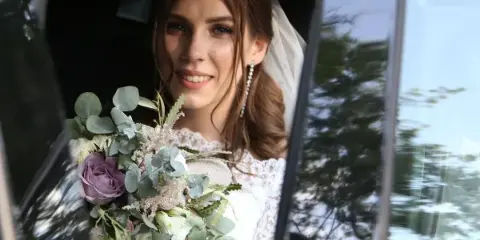 A bride in a white lace wedding dress and veil holding a floral bouquet while seated in a car.
