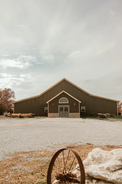 Exterior view of a large brown barn wedding venue with a gravel driveway and rustic wagon wheel in the foreground.