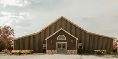 Exterior view of a large brown barn wedding venue with a gravel driveway and rustic wagon wheel in the foreground.