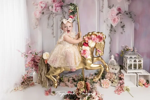 A young child in a white dress sitting on a golden rocking horse prop in a floral studio setting, watermarked with Taylored Images.