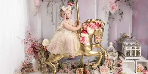 A young child in a white dress sitting on a golden rocking horse prop in a floral studio setting, watermarked with Taylored Images.