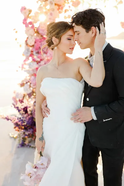 A bride in a white strapless gown and a groom in a black suit standing close together in front of a floral arch.