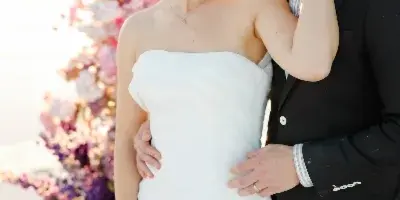 A bride in a white strapless gown and a groom in a black suit standing close together in front of a floral arch.