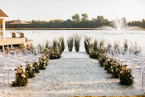 A wedding ceremony aisle decorated with floral arrangements and greenery leading to a water feature.