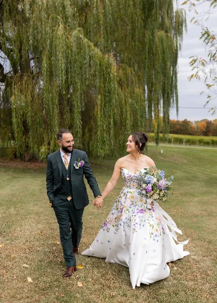 A bride in a floral wedding dress and a groom in a dark suit walking hand-in-hand on a lawn near a weeping willow tree.