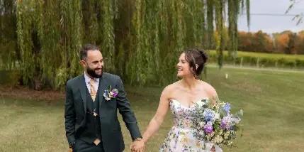 A bride in a floral wedding dress and a groom in a dark suit walking hand-in-hand on a lawn near a weeping willow tree.