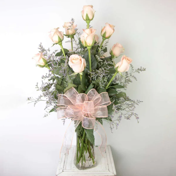 A floral arrangement featuring pale pink roses and greenery in a clear glass vase, accented with a large pink ribbon bow.