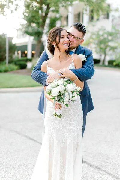 A bride in a white lace gown and a groom in a blue suit embrace while holding a white bouquet.