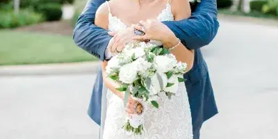 A bride in a white lace gown and a groom in a blue suit embrace while holding a white bouquet.