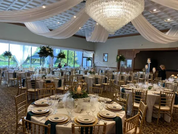 A formal wedding reception setup in a banquet hall featuring round tables with white linens, gold chiavari chairs, and a large crystal chandelier.