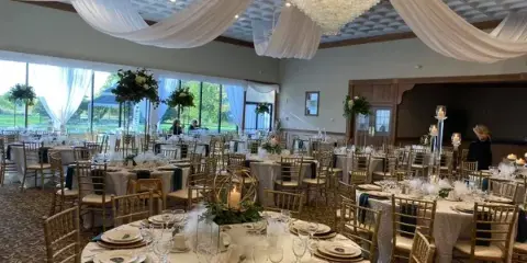 A formal wedding reception setup in a banquet hall featuring round tables with white linens, gold chiavari chairs, and a large crystal chandelier.