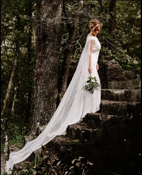 Bride in white wedding dress and long veil holding a bouquet on stone steps among trees at a historic outdoor ruins venue.
