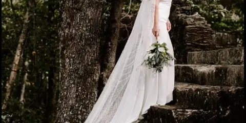 Bride in white wedding dress and long veil holding a bouquet on stone steps among trees at a historic outdoor ruins venue.