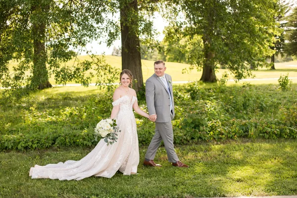 A bride and groom walking hand-in-hand on a grassy lawn under trees.