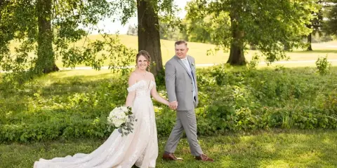 A bride and groom walking hand-in-hand on a grassy lawn under trees.
